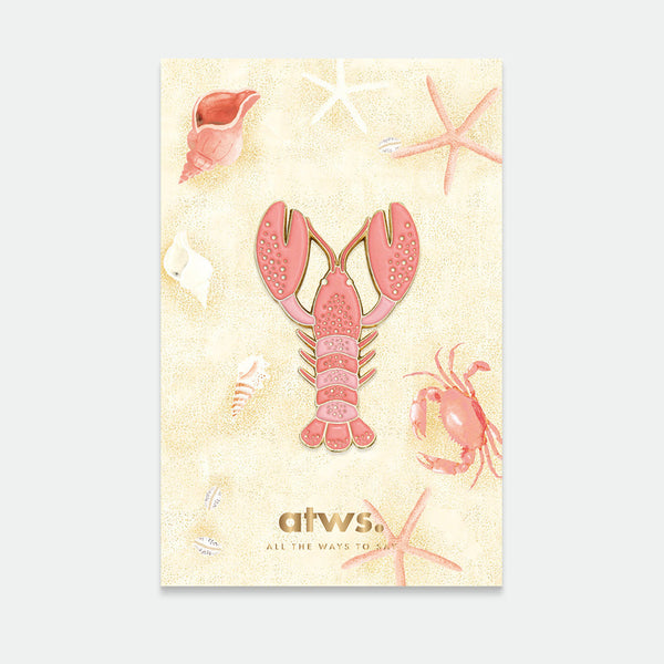 A card with a gold-plated enamel pin depicting a lobster, surrounded by starfish and shells on a beige background.