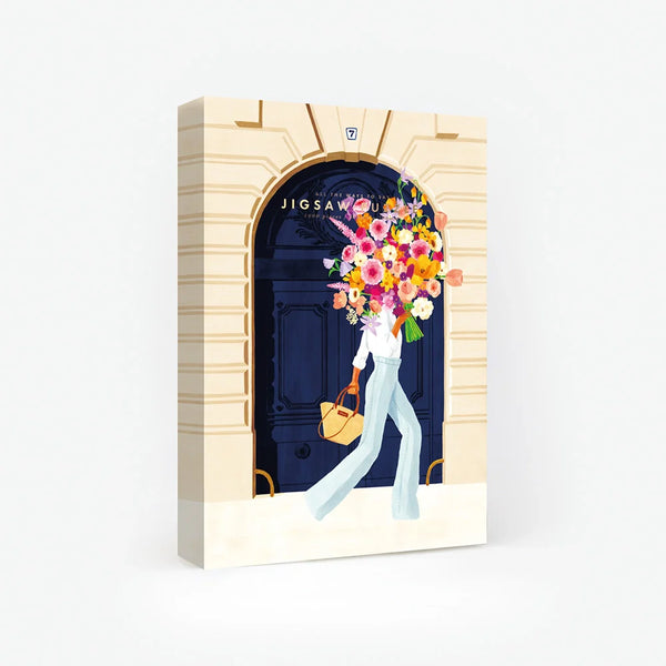 1000-piece jigsaw puzzle box by All The Ways To Say featuring a woman carrying a massive floral bouquet in front of a grand navy blue Parisian door.