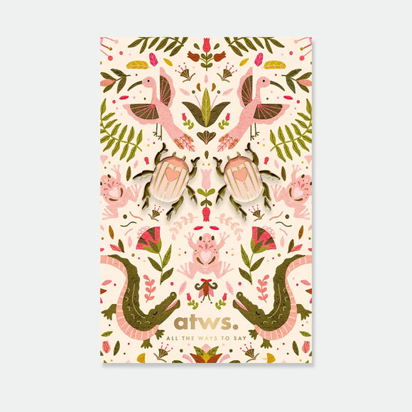 A set of two symmetrical pink beetle enamel pins with gold hearts on their backs, displayed on a lush botanical backing card.