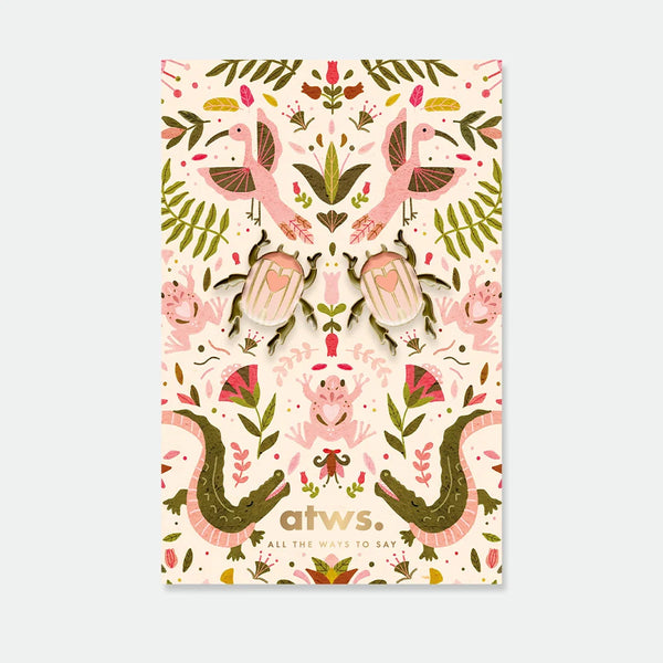 A set of two symmetrical pink beetle enamel pins with gold hearts on their backs, displayed on a lush botanical backing card.