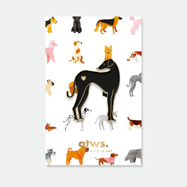 A sleek black greyhound enamel pin by All The Ways To Say with a small gold heart, displayed on a dog-themed grid backing card.