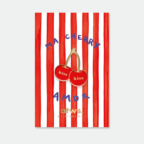 A gold enamel pin by All The Ways To Say featuring two red cherries with the word "kiss" on them, displayed on a "Ma Cherry Amor" striped card.