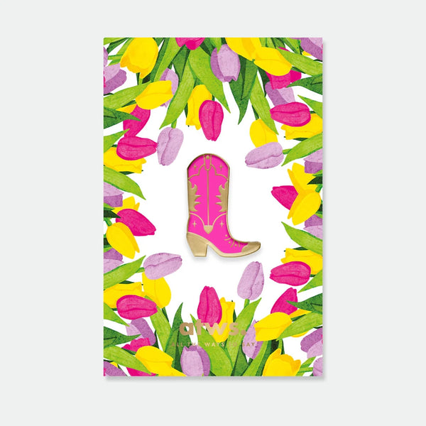 A bright pink cowboy boot enamel pin by All The Ways To Say with gold details, displayed on a colorful tulip wreath backing card.