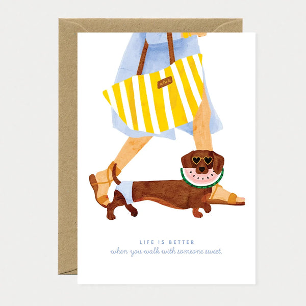 A greeting card by All The Ways To Say featuring a watercolor illustration of a person walking a dachshund wearing heart-shaped sunglasses and a watermelon collar.