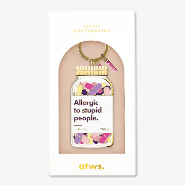 Un porte-clés humoristique en émail doré All The Ways To Say en forme de flacon de pilules avec le texte "Allergic to stupid people.", présenté dans son étui.