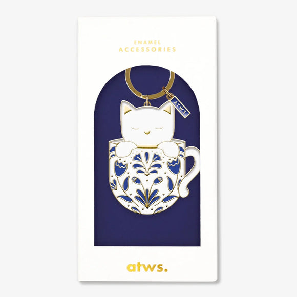 A cute gold enamel keychain by All The Ways To Say featuring a white cat in a blue patterned mug, presented in its navy blue gift box.