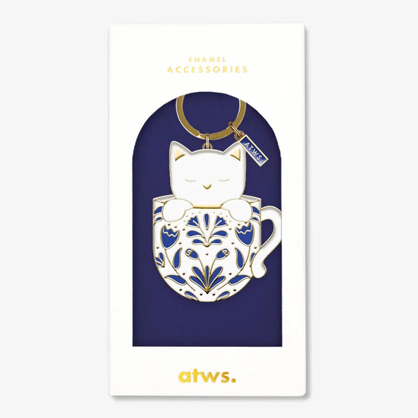 A cute gold enamel keychain by All The Ways To Say featuring a white cat in a blue patterned mug, presented in its navy blue gift box.