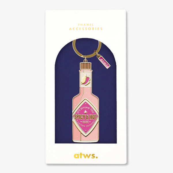 A bold gold enamel keychain by All The Ways To Say shaped like a pink hot sauce bottle with the text "IT'S GIVING YOU SPICY & HOT - BABE", in its gift box.