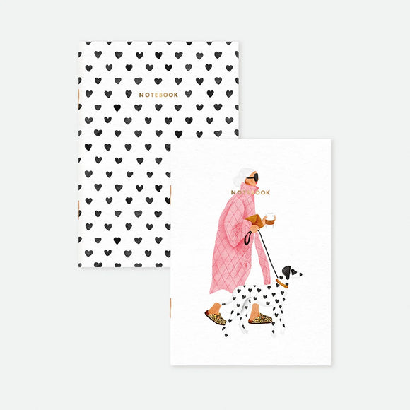 A set of two A6 pocket notebooks by All The Ways To Say: one with a black heart pattern and gold foil, the other featuring a woman walking a Dalmatian.