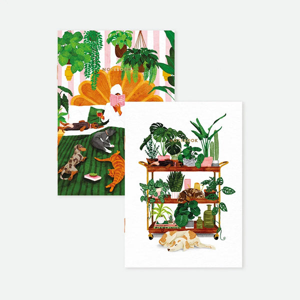 A set of two A6 pocket notebooks by All The Ways To Say: one featuring a cozy bedroom scene with pets and plants, the other a white cover with a botanical plant cart and sleeping dogs, both with gold foil.