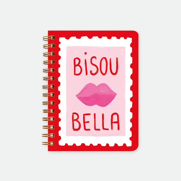 A large red spiral notebook by All The Ways To Say featuring a pink stamp with lips, the text "BISOU BELLA", and gold foil details.