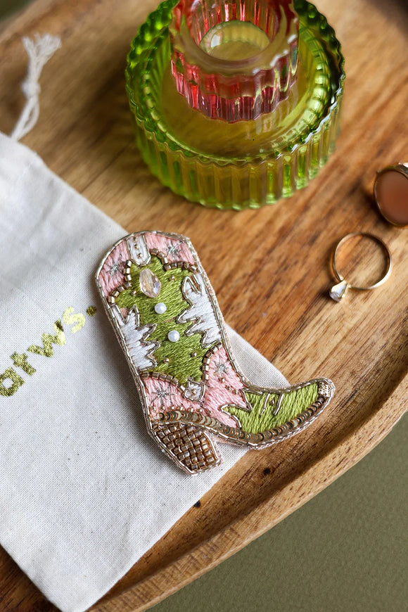 Top view of a western-style boot pin featuring intricate beadwork and embroidery on a wooden tray next to a cotton pouch.