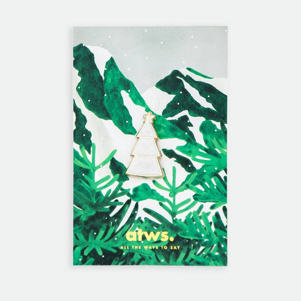 White Christmas Tree enamel pin by All the Ways to Say featuring gold-plated white tree with star detail on illustrated snowy mountain background with green pine branches.