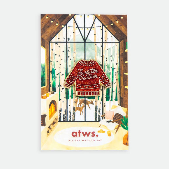 Sweater Weather enamel pin by All the Ways to Say featuring red glitter sweater with gold-plated details on illustrated winter cabin background with fireplace and forest view.