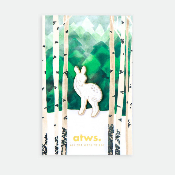 Bébé biche enamel pin by All the Ways to Say featuring gold-plated deer design on illustrated green forest background with birch trees and mountains.