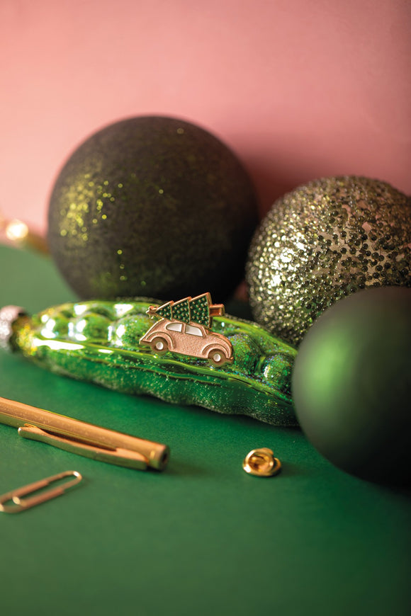 Christmas car enamel pin, a pink vintage car carrying a Christmas tree on its roof, among green glitter baubles