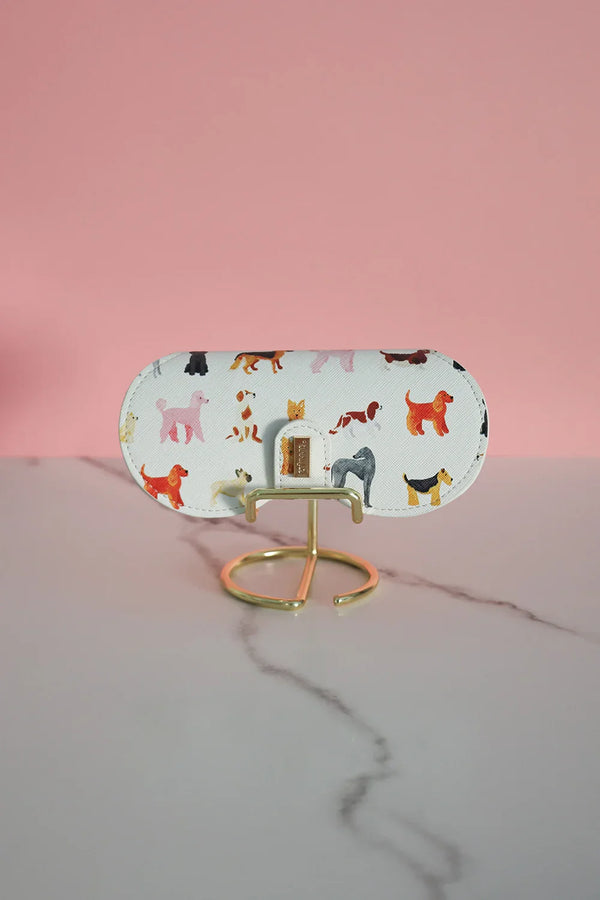 White glasses case with colorful dog illustrations, displayed on a gold stand against a pink background.