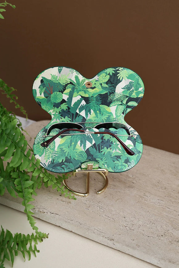 Open Green Cliff sunglasses case showing interior tropical leaves print, holding sunglasses, placed on marble stand with fern decor.