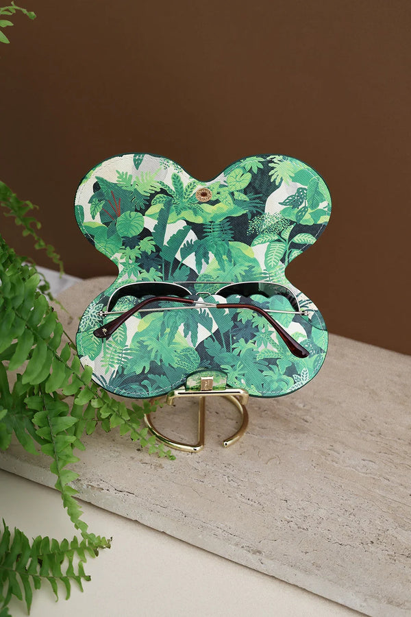 Open Green Cliff sunglasses case showing interior tropical leaves print, holding sunglasses, placed on marble stand with fern decor.