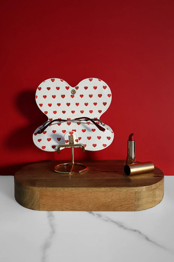 White glasses case with red heart pattern opened on a wooden stand, displayed with a red lipstick against a deep red background.