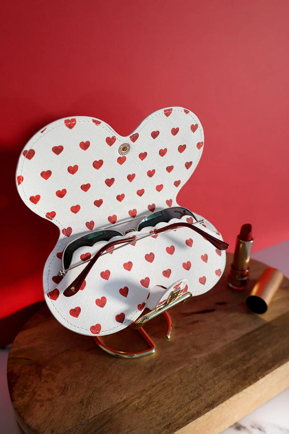 Close-up of a white glasses case with red hearts holding sunglasses, placed on a wooden stand next to a red lipstick, with a bold red backdrop.