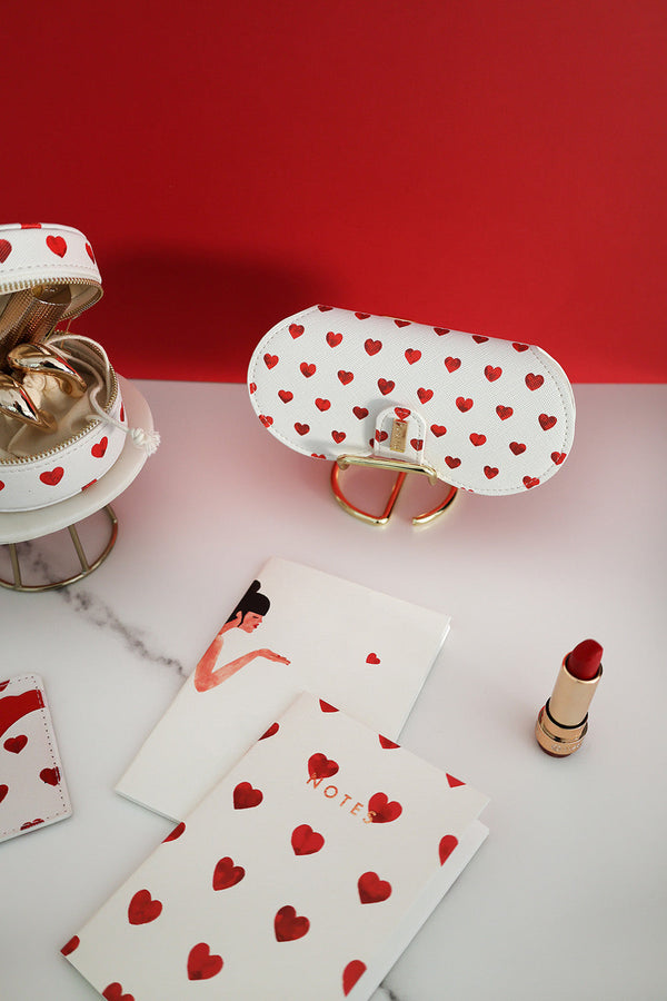 Lifestyle shot of red hearts glasses case with matching accessories, notebooks and lipstick on marble surface