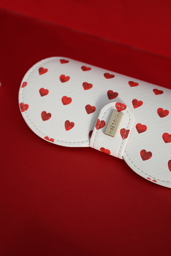 White glasses case decorated with red hearts pattern and gold logo plate against a red backdrop
