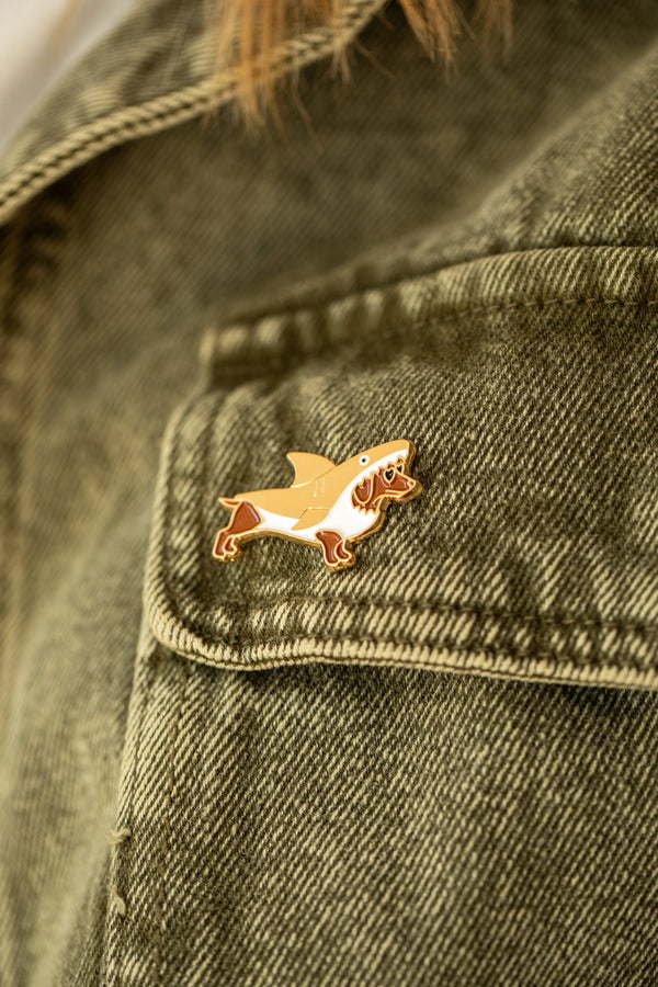 Shark Attack enamel pin, a dachshund in a shark costume with heart sunglasses, pinned on an olive denim jacket pocket