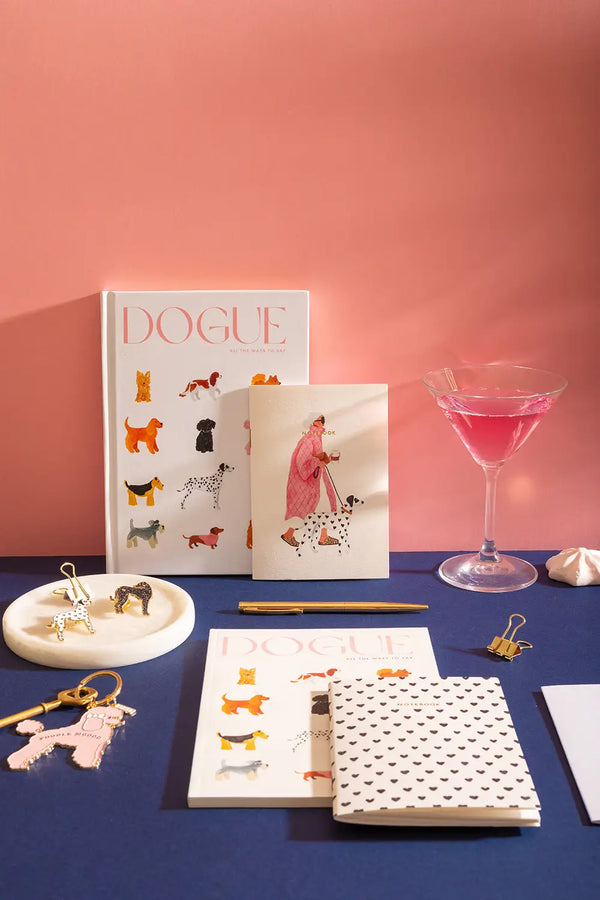 DOGUE - Collection: a stylish flat lay featuring "Dogue" notebooks, enamel pins, and a pink cocktail.