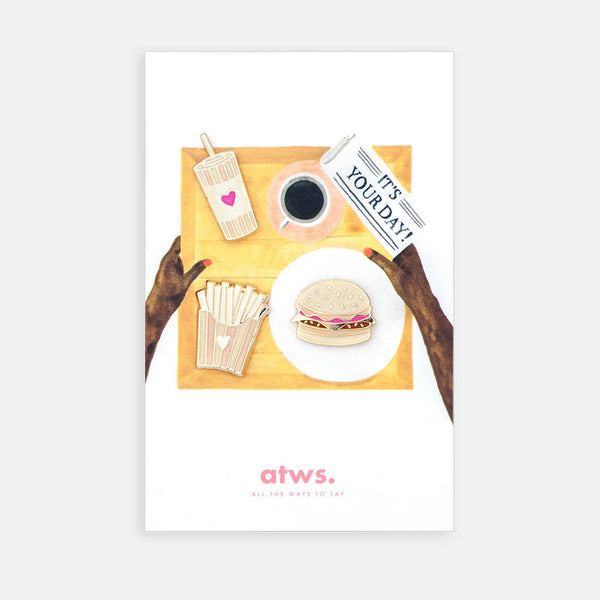 Junk Food XL enamel pin by All the Ways to Say featuring gold-plated burger, fries and drink design on illustrated tray background with newspaper and coffee.