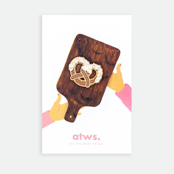 Bretzel enamel pin by All the Ways to Say featuring gold-plated pretzel with white enamel detail on illustrated cutting board background.