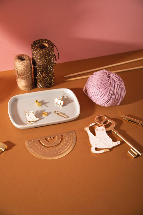 White cat enamel pin on a ceramic tray next to the matching "Don't give a sh*t" cat keychain, with yarn and knitting needles
