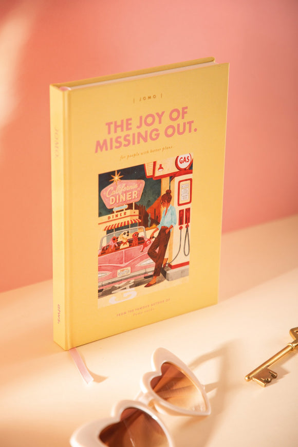Jomo A5 hardcover notebook standing at an angle, showing the yellow cover, illustrated gas station scene and pink ribbon bookmark