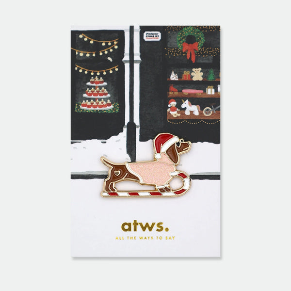 Enamel pin of a dachshund shaped like a candy cane sleigh, wearing a Santa hat, with a heart detail