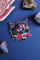 Hand-embroidered Mexican tiger brooch with three eyes, pink tongue, and gold thread details on a deep blue background with crystals.