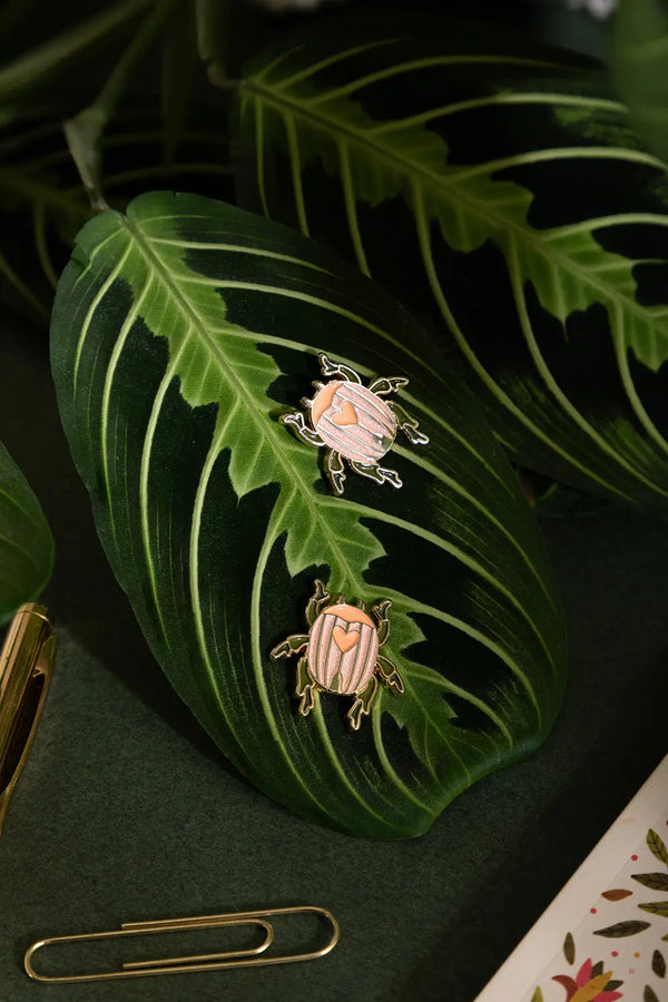 Duo Scarabée - Pins: two pink beetle enamel pins styled on a vibrant green tropical leaf.