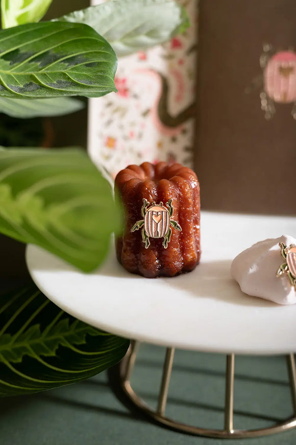 Duo Scarabée - Pins: a pink enamel beetle pin with a heart detail displayed on a French canelé pastry.