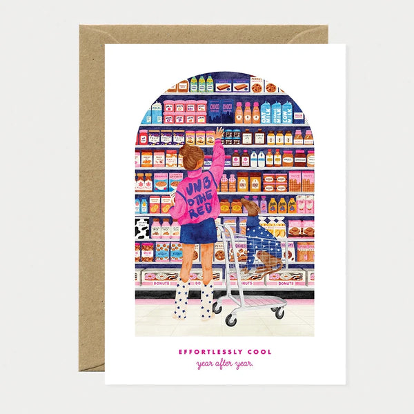 Illustrated birthday greeting card of a woman in a pink jacket reaching for a shelf in a candy and grocery aisle, with a dachshund sitting in the shopping cart below, text reading "Effortlessly cool, year after year"