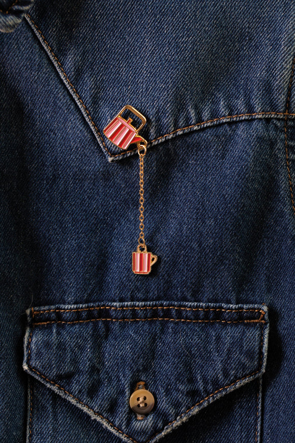 Tea Time enamel pin, a striped red and pink teapot linked by a gold chain to a matching cup, worn on a dark denim jacket