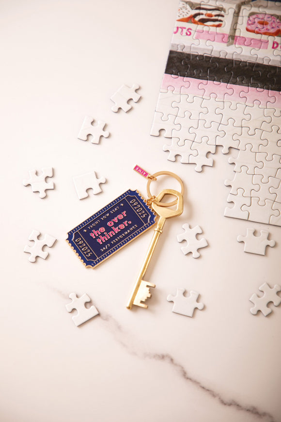 Navy "The overthinker" enamel keychain shaped like a ticket, reading "Front row seat, 24/7 performance", with a gold key and puzzle pieces
