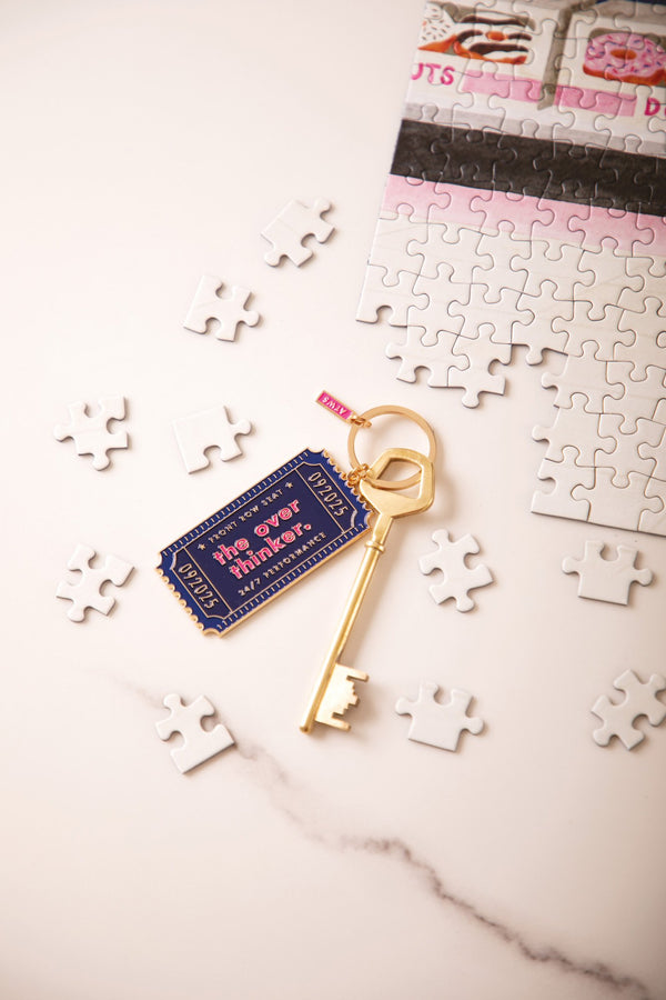 Navy "The overthinker" enamel keychain shaped like a ticket, reading "Front row seat, 24/7 performance", with a gold key and puzzle pieces